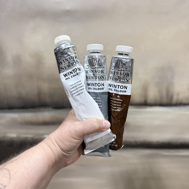 Jen holding Winsor & Newton Winton oil tubes in Titanium White, Payne's Gray and Vandyke Brown in front of her canvas